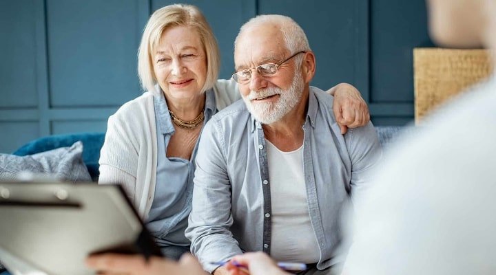 Older man and woman seated closely on a sofa, leaning toward a consultant holding a clipboard in the foreground; they appear attentive and engaged. Interior office or living-room setting with a blue paneled wall and cushions. No visible text. Calm, reassuring tone.